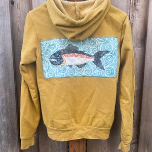 May include: A mustard yellow hoodie with a zipper closure and a large embroidered patch on the back. The patch features a fish swimming in a blue and white swirling pattern.
