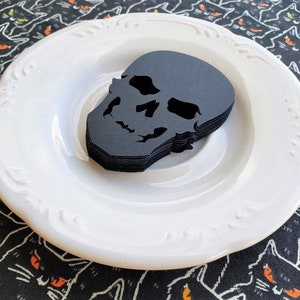 May include: A white plate with a stack of black paper skull cutouts on top. The plate is on a black and white fabric with a pattern of black cats with orange eyes.