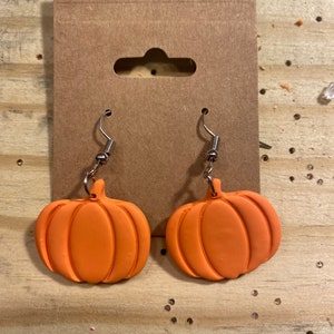 Puede incluir: Un par de pendientes de calabaza naranja con ganchos plateados. Los pendientes est&aacute;n hechos de arcilla polim&eacute;rica y tienen una superficie texturizada.