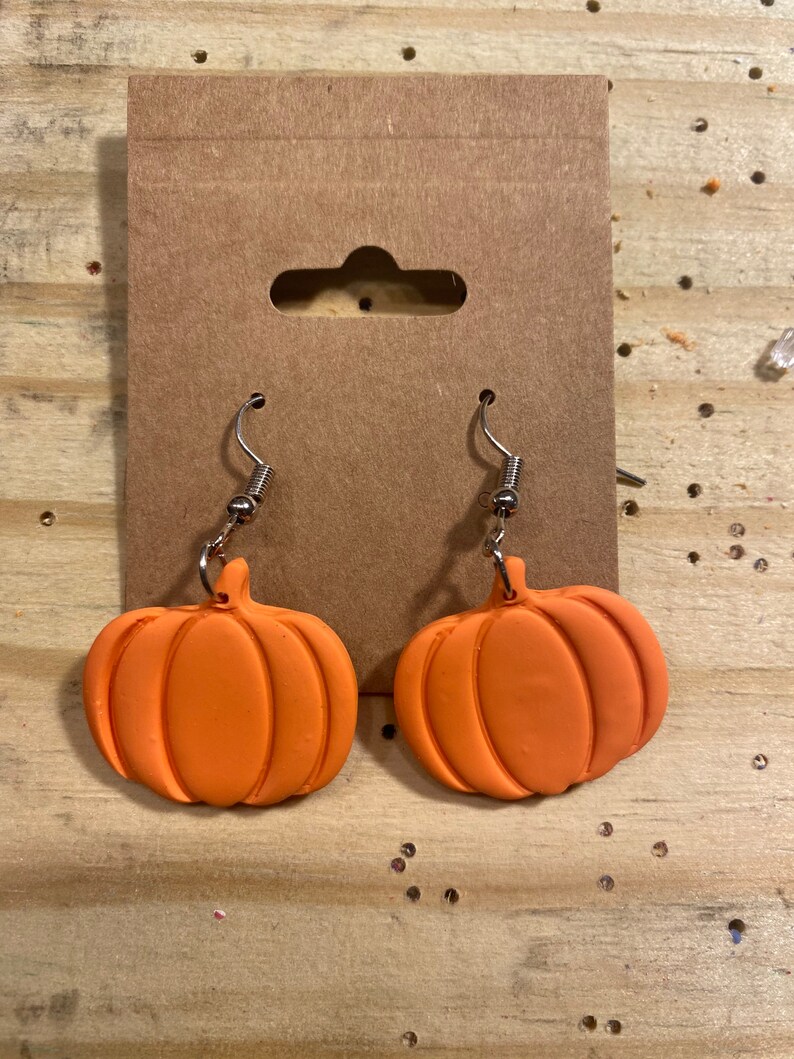 Puede incluir: Un par de pendientes de calabaza naranja con ganchos plateados. Los pendientes est&aacute;n hechos de arcilla polim&eacute;rica y tienen una superficie texturizada.