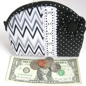 May include: A black and white chevron patterned zippered pouch with a white lace trim and a black polka dot fabric with a black fabric flower. The pouch is sitting on a dollar bill with some coins.