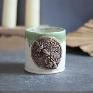 May include: A ceramic mug with a speckled white base and a green glazed rim. The front features a circular, dark brown relief medallion depicting two bees surrounded by intricate floral and leaf patterns with a rope-like border detail.