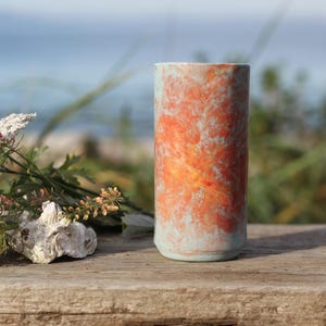 May include: A cylindrical ceramic tumbler featuring a marbled finish with blended shades of light blue, vibrant orange, and yellow. The vessel has a smooth, straight-sided profile with a flat base, showcasing a unique, swirling color pattern.