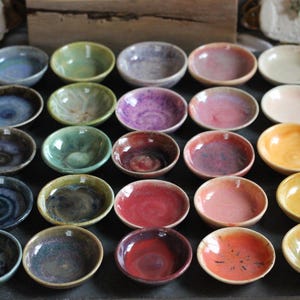 May include: A collection of small, handmade ceramic bowls in various colors, including blue, green, purple, pink, and yellow. The bowls are arranged on a dark surface, showcasing their unique glaze finishes and textures. Ideal for serving dips or small portions.