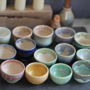 May include: A collection of small, handcrafted ceramic bowls in various colors, including blue, green, yellow, and pink. Each bowl has a unique glaze and rim design, showcasing a rustic aesthetic. The bowls are arranged on a dark surface.