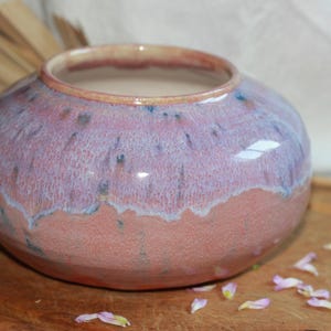 May include: A round ceramic vase with a wide opening. The vase has a two-tone color scheme, with a reddish-orange base and a lavender-purple upper section. Small blue speckles dot the surface. The vase sits on a wooden surface.