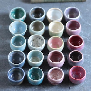 May include: A collection of twenty small, round ceramic cups arranged in a five-by-four grid. The cups feature a variety of glazed finishes in shades of blue, green, white, pink, purple, and red, each with unique color variations and textures.