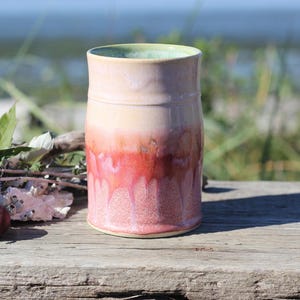 May include: A cylindrical ceramic tumbler featuring a smooth, light cream-colored upper section that transitions into a vibrant, textured coral-pink base with a distinct drip glaze effect that runs vertically down the exterior surface of the vessel.