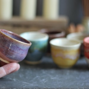 May include: A collection of small, handmade ceramic cups in various colors. The cups are displayed on a dark surface, with one cup in the foreground held by a hand. The cups have a rustic, artisanal appearance, with a focus on the unique glaze and shape.