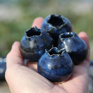 May include: A collection of four small, round, dark blue ceramic vessels with a glossy finish and uneven, ruffled openings. Each piece features a deep indigo glaze with subtle variations in tone, creating a smooth, reflective surface texture.