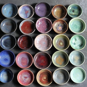 May include: A collection of twenty-five small, round ceramic bowls arranged in a five-by-five grid. Each bowl features a unique reactive glaze finish in various colors, including shades of blue, red, purple, green, brown, and cream.