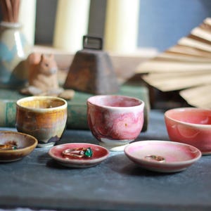 May include: A collection of handmade ceramic pieces, including small bowls and ring dishes, in shades of brown, pink, and yellow. The items are arranged on a dark surface, with a blurred background featuring candles and decorative objects.