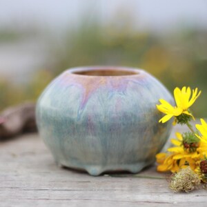 May include: A small, round ceramic vase with a light blue and iridescent glaze. The vase has a wide opening and sits on three small feet. Bright yellow flowers and greenery are arranged beside the vase on a wooden surface.