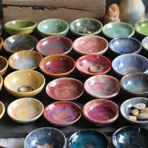 May include: A collection of small, handmade ceramic bowls in various colors, including green, blue, yellow, and red. Some bowls contain rings or small stones. The bowls are arranged on a dark surface, showcasing their unique textures and glazes.