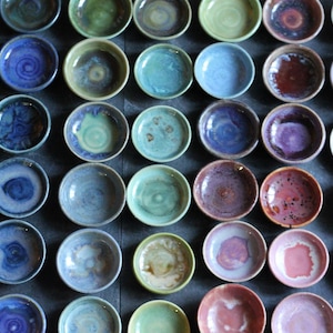 May include: A collection of small, round ceramic bowls arranged in a grid. Each bowl features a unique glazed finish in various colors, including shades of blue, green, purple, pink, and brown. The surfaces show variations in color intensity.