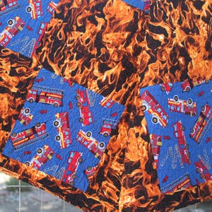 May include: A quilted blanket featuring a vibrant flame pattern in shades of orange, yellow, and black. Accented with patches of blue fabric adorned with red fire trucks. The blanket is designed with a patchwork style.
