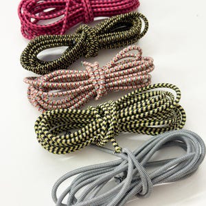 May include: Five coiled lengths of elastic shock cord arranged in a vertical row. The cords feature distinct color patterns including solid pink with black, black with yellow, multi-colored, yellow with black, and grey with white striped designs.