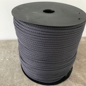 May include: A large spool of dark gray cord featuring a tightly woven, braided texture. The cord is wound neatly around a black plastic cylindrical reel with a circular top and bottom, creating a uniform and dense stack of gray material.