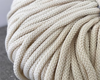 Puede incluir: Primer plano de una gran bobina de cordón de algodón grueso de color crema. El cordón está enrollado apretado y tiene un acabado liso y ligeramente brillante.