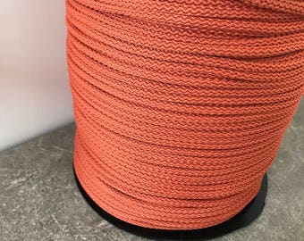 Puede incluir: Una bobina de cordón de algodón naranja enrollada alrededor de una bobina de plástico negro. El cordón está enrollado apretado y tiene una superficie texturizada.