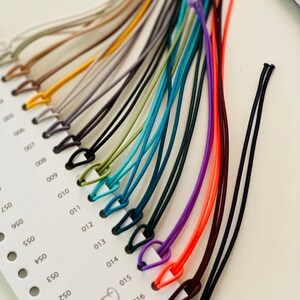 May include: A color chart featuring a variety of thin, colorful cords with small loops. The cords range in color from neutral tones to vibrant hues like orange, purple, and teal. The chart is labeled with numbers and the brand name "DeLuna".