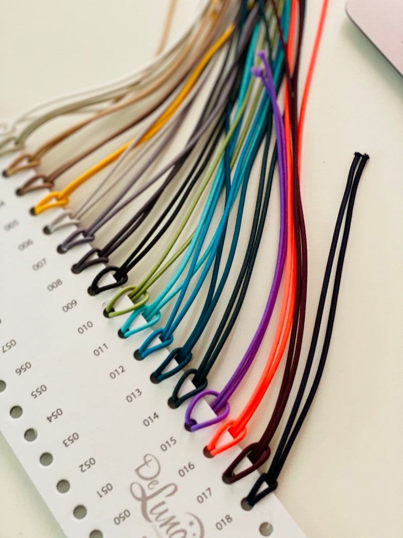 May include: A color chart featuring a variety of thin, colorful cords with small loops. The cords range in color from neutral tones to vibrant hues like orange, purple, and teal. The chart is labeled with numbers and the brand name "DeLuna".