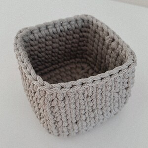 May include: A square-shaped, crocheted storage basket made of beige yarn. The basket is empty and has a textured surface.