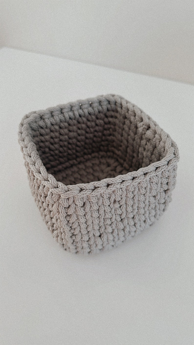 May include: A square-shaped, crocheted storage basket made of beige yarn. The basket is empty and has a textured surface.