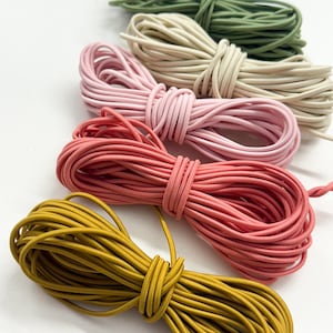 May include: Five bundles of elastic cord arranged in a diagonal row. The colors from top to bottom are sage green, cream, light pink, coral pink, and mustard yellow. Each bundle is neatly coiled and secured with a loop of the same cord material.
