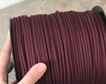May include: A spool of burgundy-colored cord. The cord is tightly wound and has a textured appearance. The spool is held by a hand, showcasing the color and texture of the cord. The cord appears to be made of a durable material.
