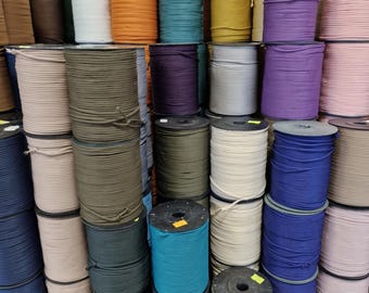 May include: A large collection of cylindrical spools stacked together, each wrapped with a continuous length of cord in various solid colors including blue, green, white, black, purple, pink, yellow, brown, and gray, creating a dense display.