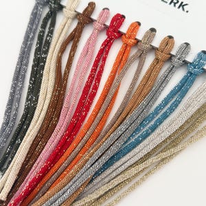 Op de afbeelding: Een collectie van twaalf metallic koorden bevestigd aan een witte displaykaart met het label NUSINERK. De koorden hebben een glinsterende, geweven textuur en zijn verkrijgbaar in diverse kleuren, waaronder grijs, bruin, crème, roze, rood, oranje en blauw.