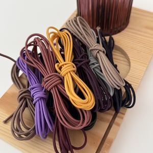 May include: A collection of elastic hair ties in various colors including brown, purple, mustard yellow, dark red, beige, grey, and black. Each hair tie is folded and secured with a knot, creating a bundle of multiple circular elastic bands.