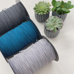 May include: Three spools of elastic cord in gray, teal, and light gray are displayed. The spools are positioned in front of three small potted succulents with green leaves. The background is a plain white surface.