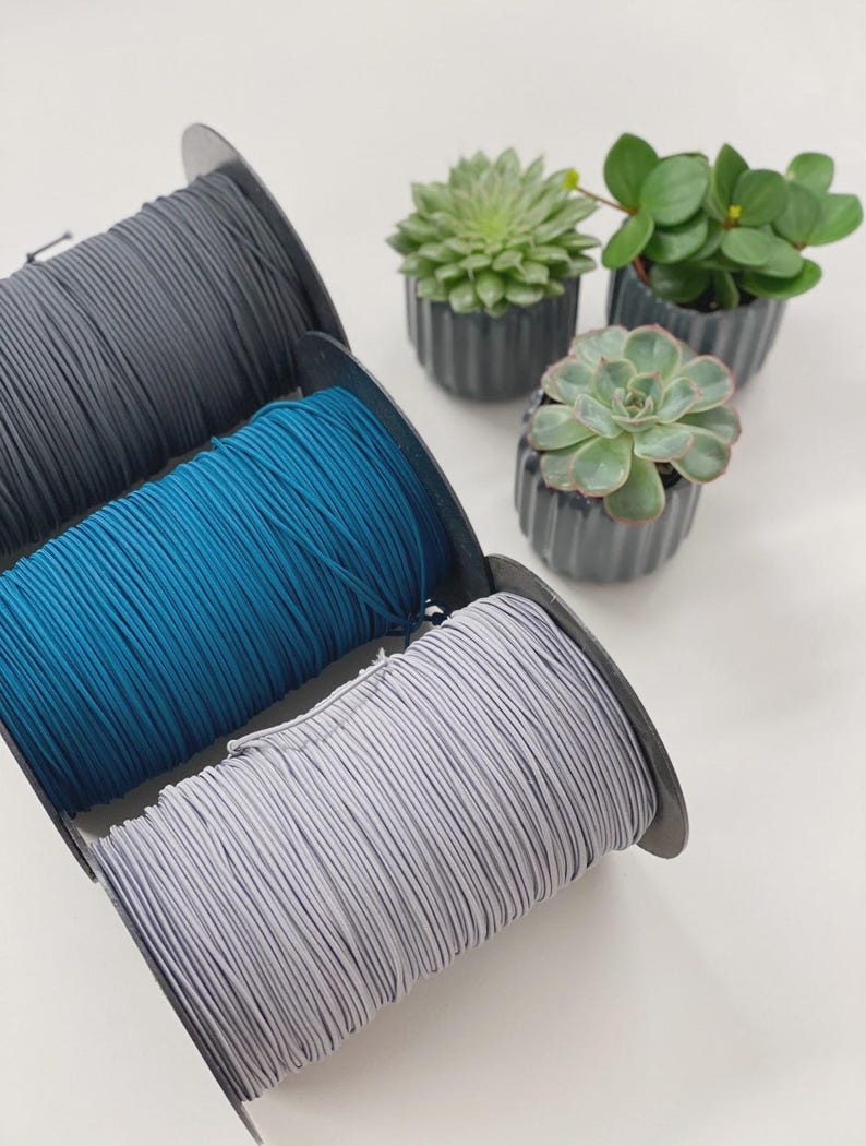 May include: Three spools of elastic cord in gray, teal, and light gray are displayed. The spools are positioned in front of three small potted succulents with green leaves. The background is a plain white surface.