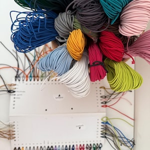 May include: A collection of colorful cords in various hues, including blue, green, yellow, and pink, are displayed in a woven basket. The cords are connected to white panels with labeled holes, suggesting a crafting or DIY project. The image showcases a variety of colors and textures.