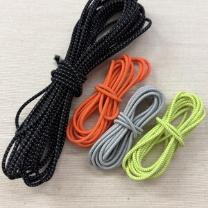 Könnte beinhalten: Vier aufgewickelte elastische Kordeln in Schwarz mit weißen Sprenkeln, einfarbigem Orange, einfarbigem Hellgrau und einfarbigem Limettengrün. Jede Kordel ist zu einer kompakten Schlaufe gewickelt und hat eine texturierte, gewebte Oberfläche.