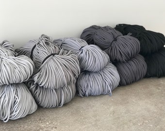 May include: A collection of gray, dark gray, and black macrame cord bundles. The cords are tightly wound and secured with black bands. The bundles are stacked against a light gray wall and a concrete floor.