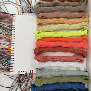 May include: A white rectangular display card with numerous holes along the side, holding a variety of colorful elastic cords. The cords are organized in horizontal bundles of different shades, including brown, tan, neon yellow, orange, pink, white, gray, green, blue, and black.