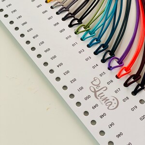 May include: A color chart featuring a variety of cords in different colors, including white, brown, gray, black, green, blue, purple, and orange. Each cord is attached to a small, triangular clasp. The chart is labeled with numbers and the "De Luna Design" logo.