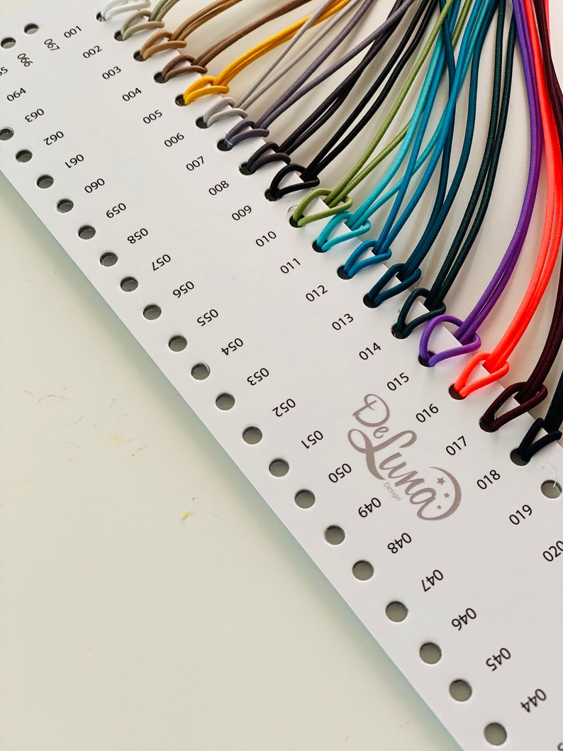May include: A color chart featuring a variety of cords in different colors, including white, brown, gray, black, green, blue, purple, and orange. Each cord is attached to a small, triangular clasp. The chart is labeled with numbers and the "De Luna Design" logo.