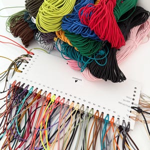 May include: A collection of multi-colored elastic cords bundled together, paired with a white display card labeled 'nusinerk' featuring numbered holes from 40 to 73. The card displays various cord samples in shades of red, blue, green, yellow, and pink.
