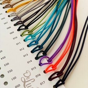 Puede incluir: Una carta de colores que muestra una variedad de cordones el&aacute;sticos en tonos beige, amarillo, gris, negro, verde, verde azulado, azul, morado, naranja y burdeos. Cada cord&oacute;n est&aacute; en bucle y sujeto a una tarjeta numerada, con el logotipo "De Luna Design".
