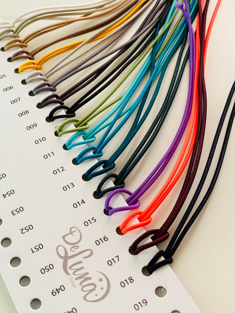 Puede incluir: Una carta de colores que muestra una variedad de cordones el&aacute;sticos en tonos beige, amarillo, gris, negro, verde, verde azulado, azul, morado, naranja y burdeos. Cada cord&oacute;n est&aacute; en bucle y sujeto a una tarjeta numerada, con el logotipo "De Luna Design".
