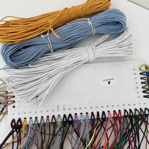 May include: Three bundles of elastic cord in mustard yellow, light blue, and white are positioned above a white display card. The card features the text nusinerk with a heart icon and displays numerous thin, multi-colored elastic cords attached.