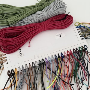 May include: A white display card featuring three large hanks of elastic cord in olive green, light gray, and dark pink. The card includes a numbered color chart from 5 to 39, with various thin, multicolored elastic cords attached to each number.