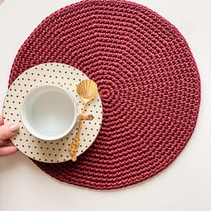 May include: A burgundy crocheted placemat with a cream-colored polka-dot plate and a gold spoon. The plate holds a white teacup. The placemat is round and has a textured surface. The spoon has a seashell design.