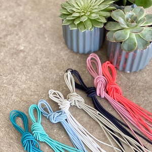 May include: A selection of colourful elastic cords, knotted at the top, in shades of teal, light blue, grey, navy, pink, and red. The cords are arranged on a textured surface, with small potted succulents in the background.