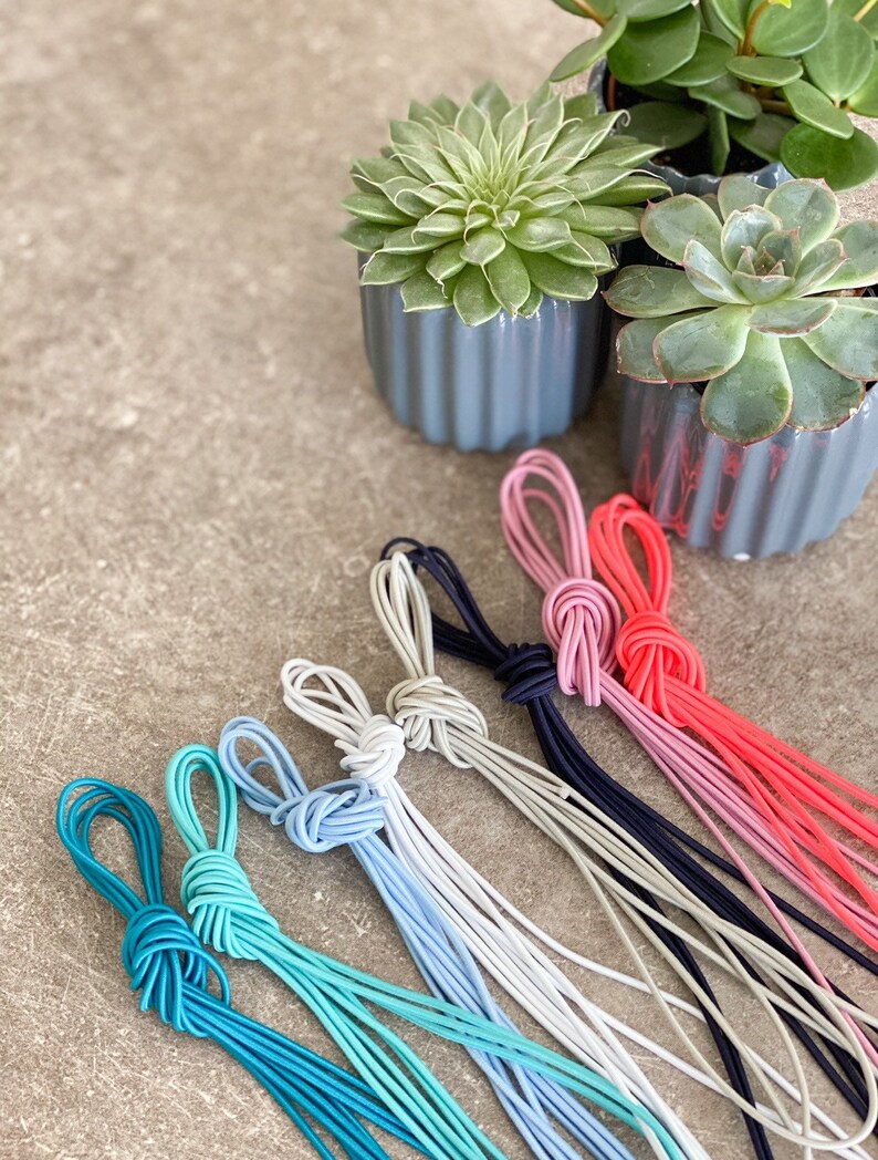 May include: A selection of colourful elastic cords, knotted at the top, in shades of teal, light blue, grey, navy, pink, and red. The cords are arranged on a textured surface, with small potted succulents in the background.