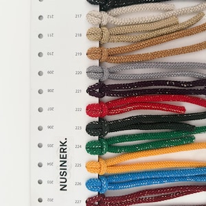 May include: A color swatch card labeled NUSINERK featuring a vertical column of numbered holes from 201 to 215 and 216 to 228. Attached to the right side are looped cords in various colors including white, black, beige, gold, gray, red, green, yellow, blue, and maroon.